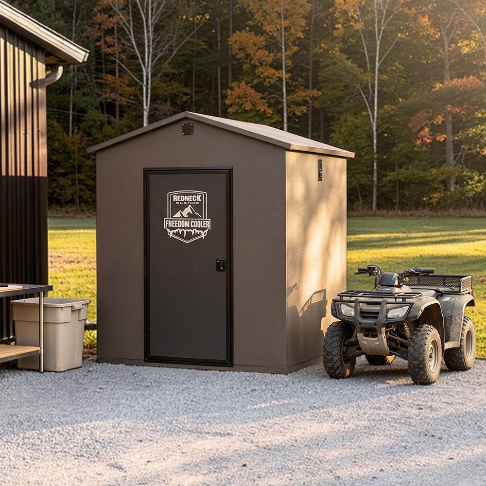 Freedom Walk-in Cooler sitting next to barn, processing table, and ATV on gravel drive, in front of a grass field with tree border.